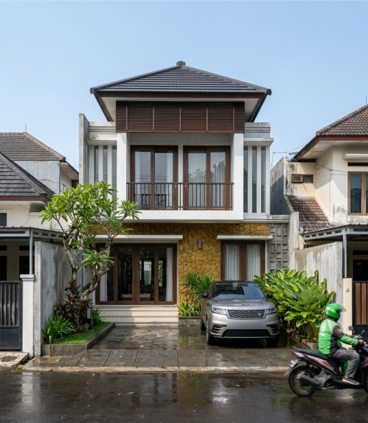 Tampilan fasad rumah minimalis tropis modern dua lantai dengan carport, balkon, batu alam, dan taman hijau.